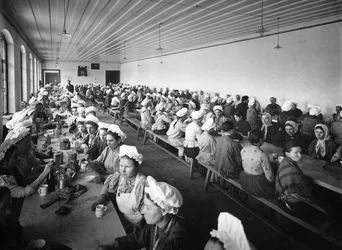 Women workers in a factory warehouse take a dinner break, supervised by a few male overseers